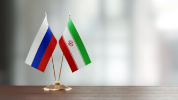 Russian And Iranian Flag Pair On A Desk Over Defocused Background