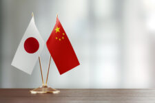 Japanese And Chinese Flag Pair On A Desk Over Defocused Background